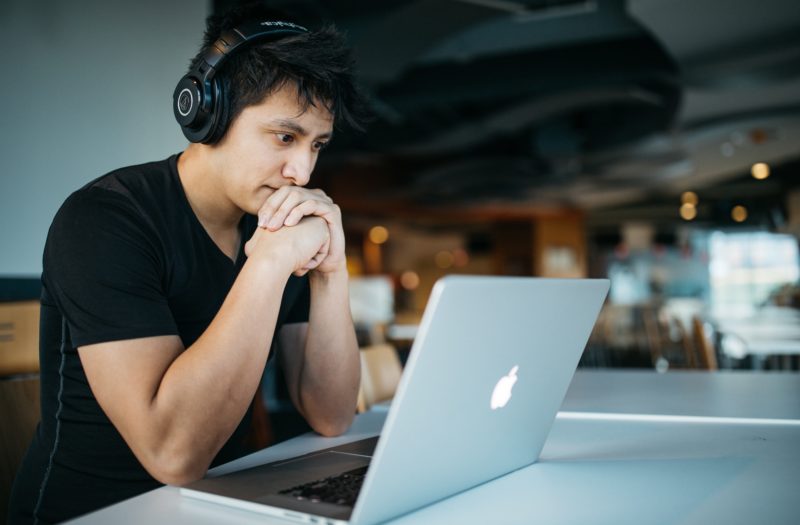 Man wearing headphones with hands folded at his mouth reviews laptop screen