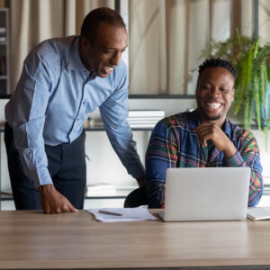 one male standing and one male sitting, while both are looking at a laptop and laughing